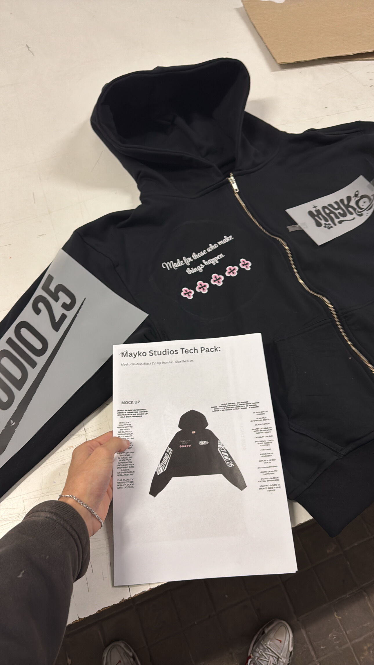 Black zip-up hoodie displayed alongside its printed tech pack specification sheet