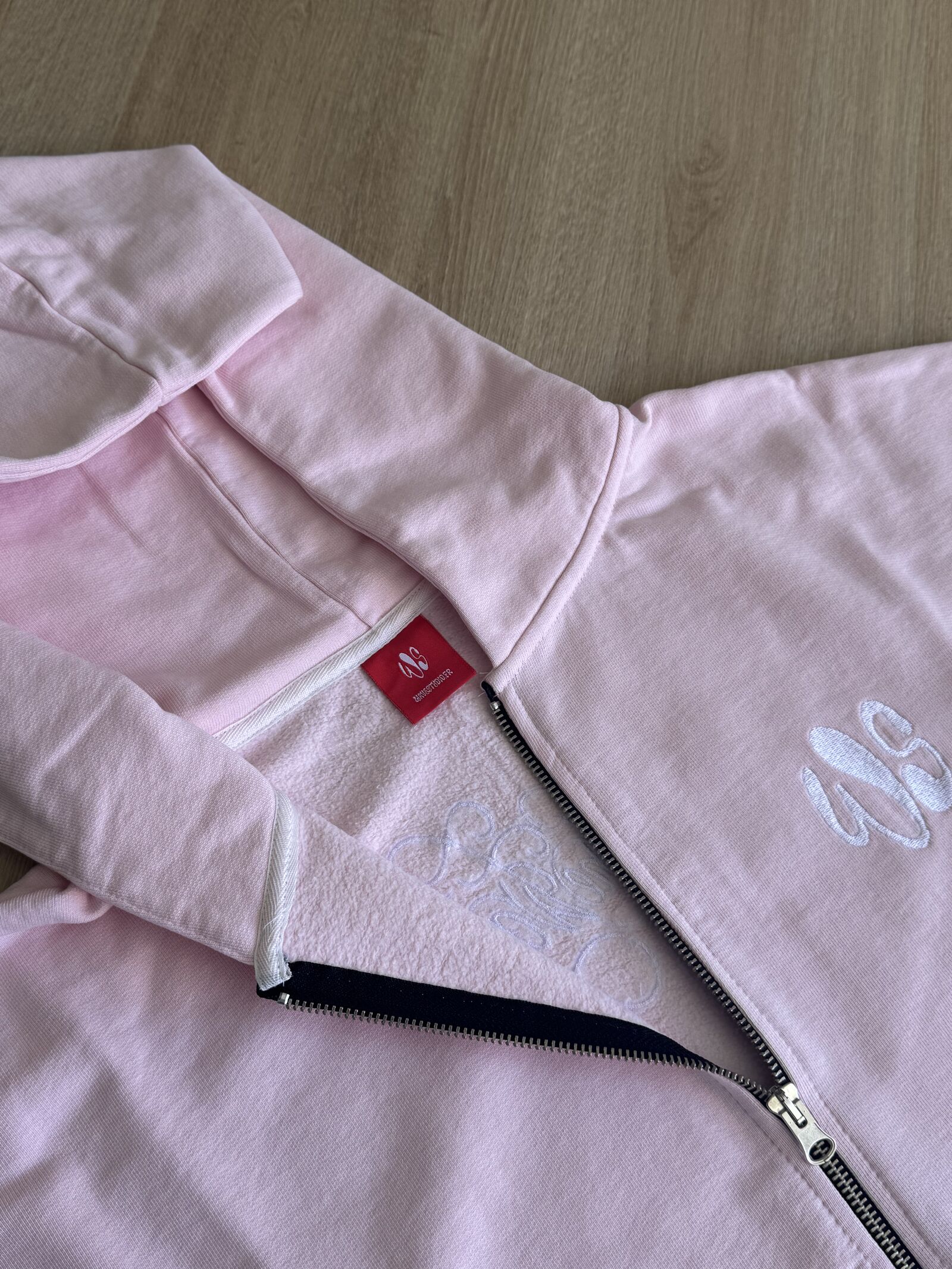 Pink zip-up hoodie with white tonal embroidered logo on wooden surface